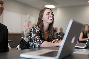A student laughing in class, with a laptop in front.